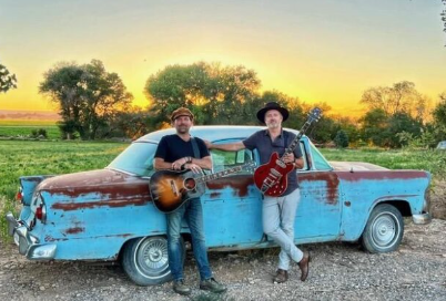 Two musicians with guitars beside a vintage turquoise car at sunset